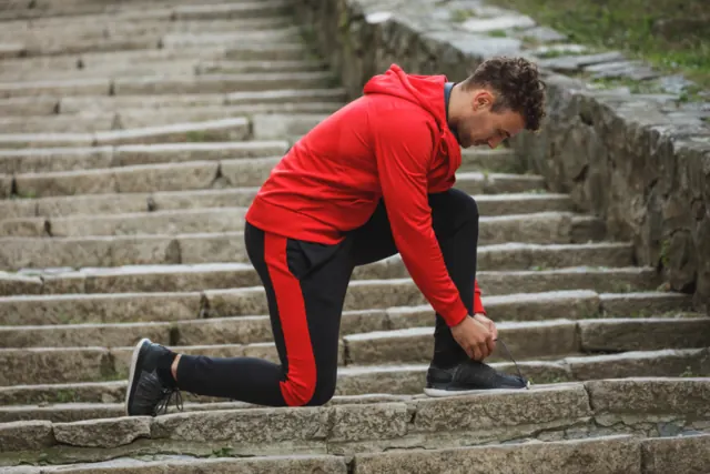 Man in a red hoodie and track pants tying his shoe on stone steps